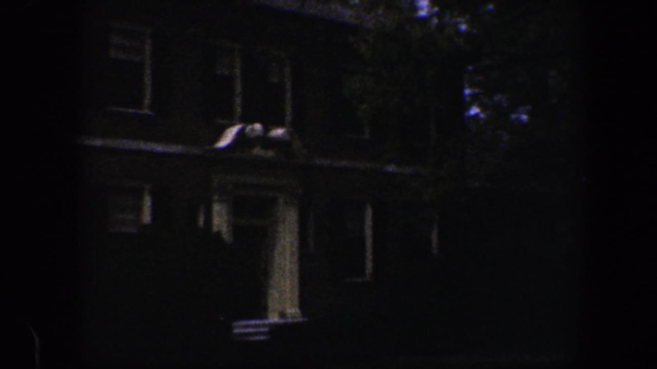 1960 Baltimore home-movie frame showing a dim house exterior near a landscaped lawn and blooming garden border.