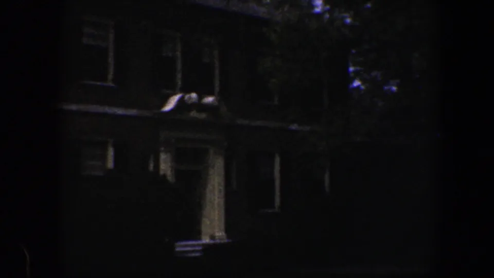 1960 Baltimore home-movie frame showing a dim house exterior near a landscaped lawn and blooming garden border.