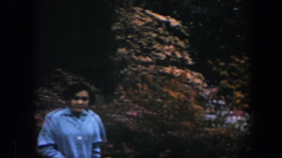 A woman in a light blue button-up shirt walks through a Baltimore garden with colorful shrubs and dark leafy background in 1960 footage.