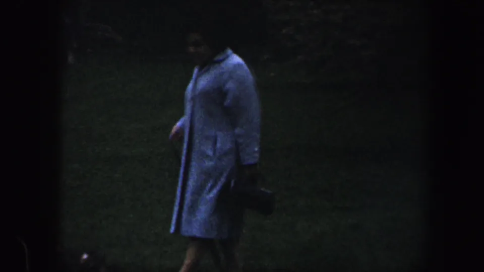 1960 Baltimore home-movie frame with a woman in a light-colored coat walking across a dim grassy yard while carrying a small dark bag.