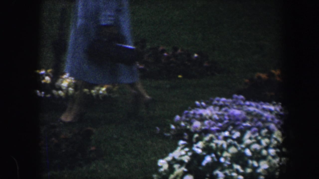 A partially seen person in a light dress moves through a 1960 Baltimore garden with flowerbeds nearby.