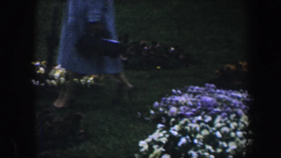 A partially seen person in a light dress moves through a 1960 Baltimore garden with flowerbeds nearby.