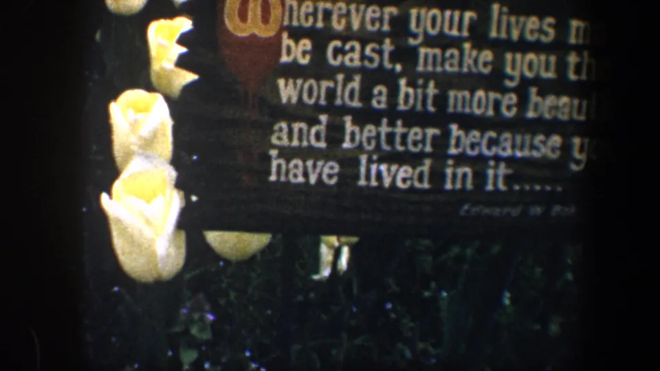 1960 Baltimore 8mm frame showing a wooden sign with white inspirational lettering beside white roses and leafy garden plants.