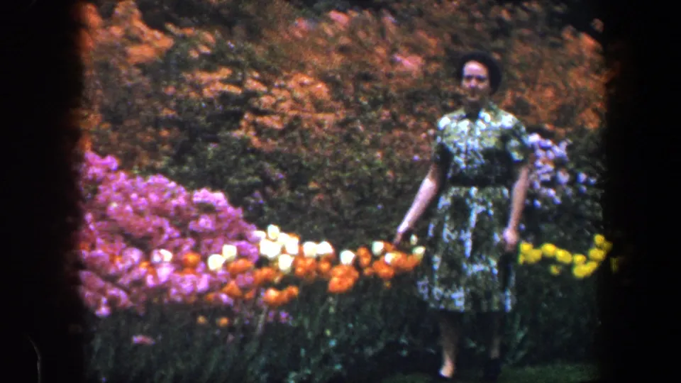 The woman faces the camera while standing near purple, white, orange, and yellow flowers in the 1960 Baltimore garden scene.
