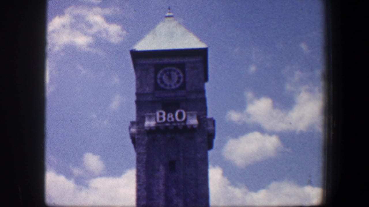 1960 Baltimore home-movie frame showing the B&O clock tower, clock face, and lettering beneath a bright clouded sky.