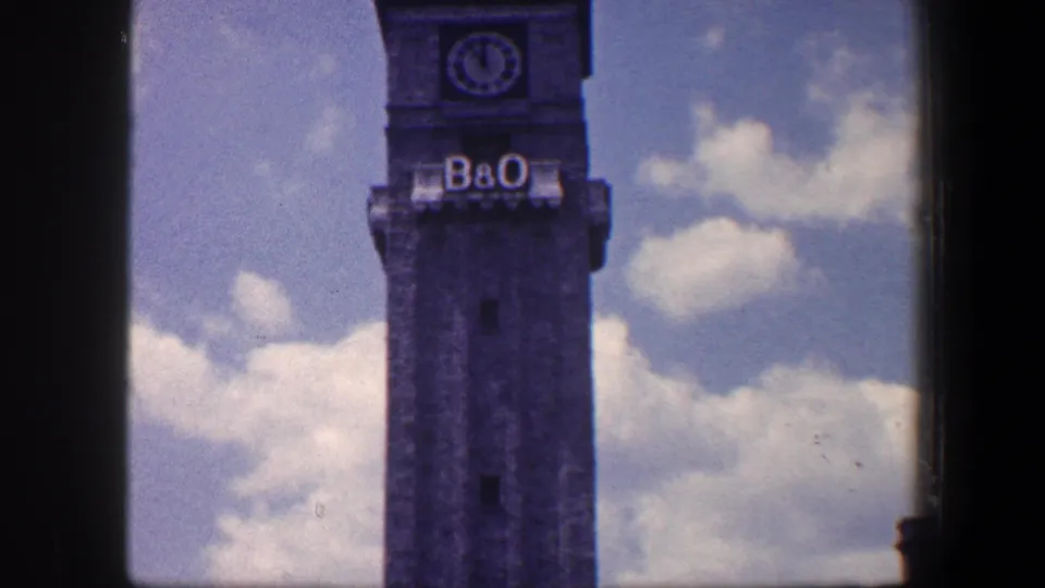 1960 Baltimore frame of the B&O clock tower from a handheld home movie, showing the tower shaft, clock face, lettering, and clouds.