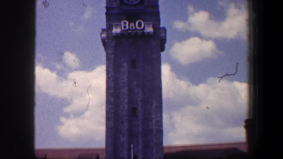 Baltimore B&O clock tower in a 1960 home-movie frame, with visible lettering, clock face, sky, clouds, and a low roofline.