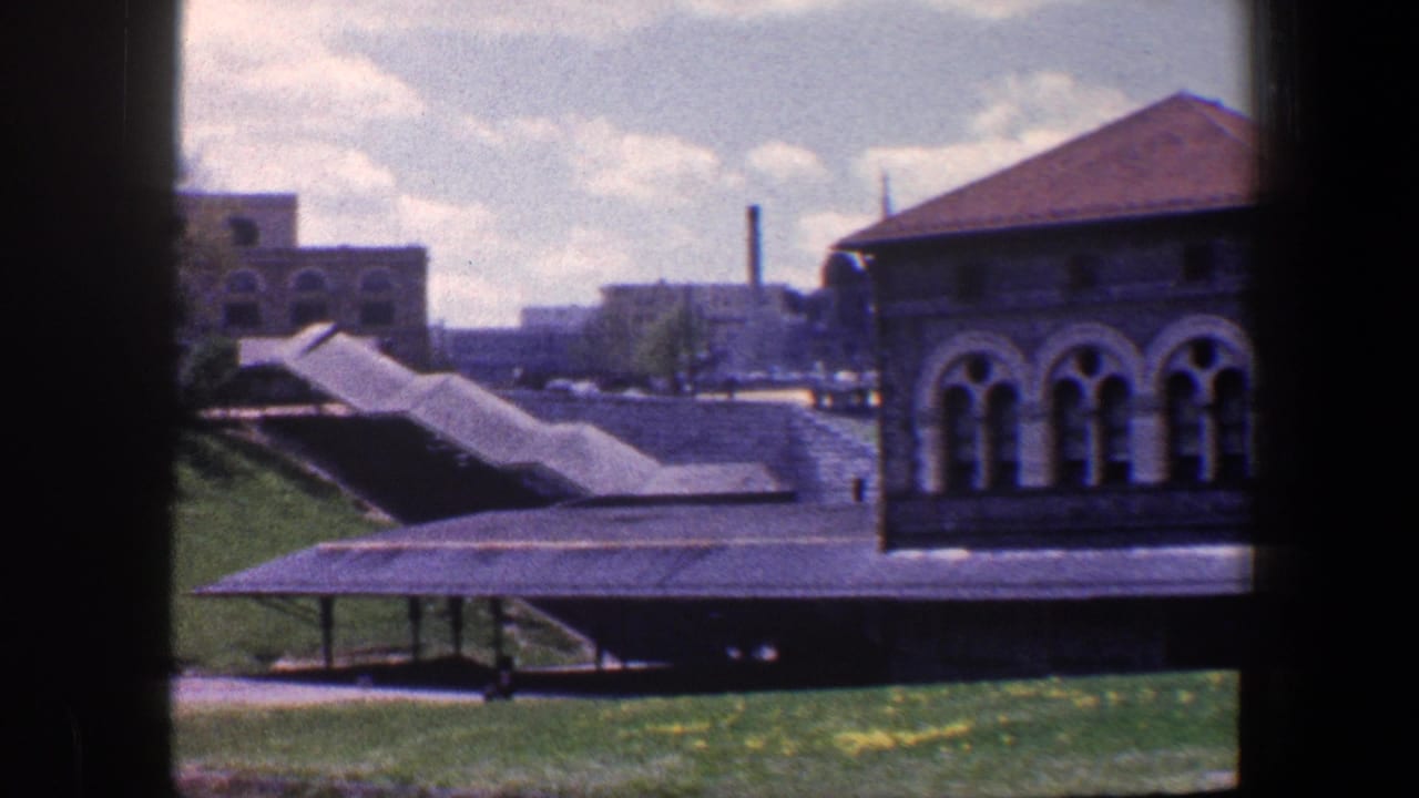 1960 Baltimore view across a covered walkway and lawn toward stone architecture with arched windows and distant industrial buildings.