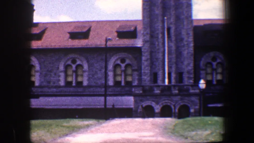 1960 Baltimore 8mm frame showing a tower-like section of the stone building, arched lower openings, windows, grass, path, and lamp post.