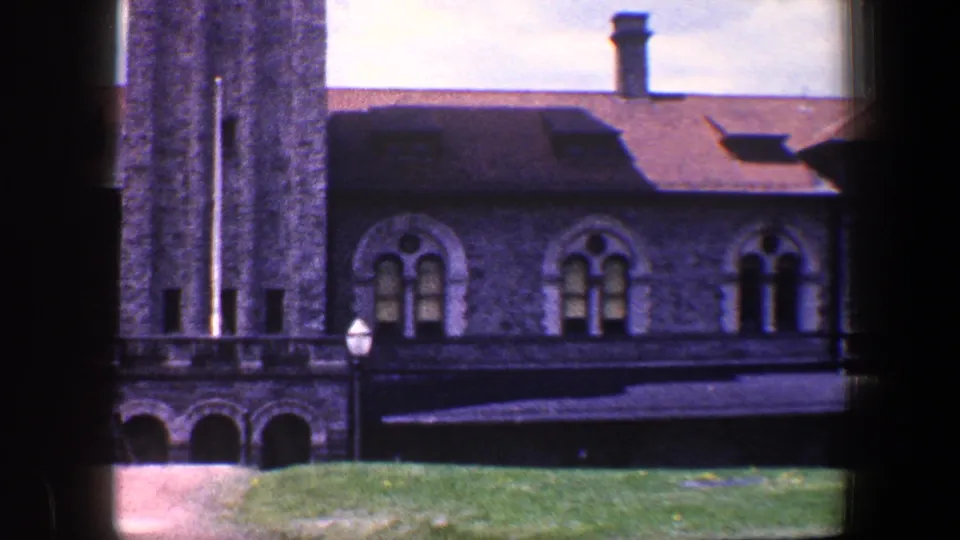 1960 Baltimore home-movie frame showing a gray stone building with arched windows, tower edge, pitched roof, lawn, and lamppost.
