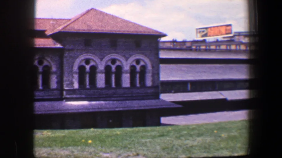 1960 Baltimore frame showing a stone building corner with arched windows, lawn, low roofline, and a distant Arrow 77 sign.