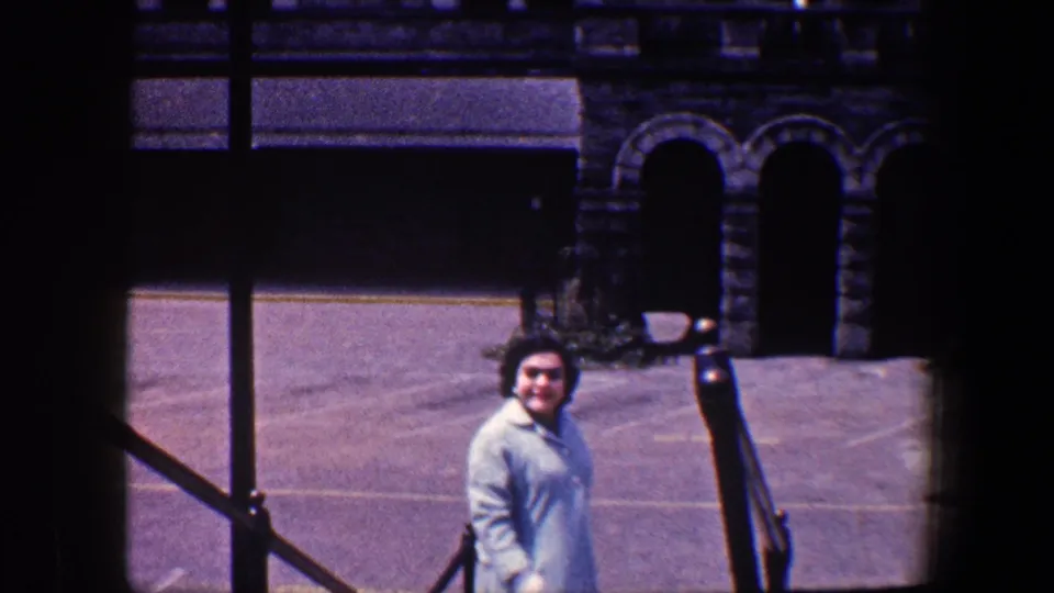 A woman in a light blue coat turns toward the camera on exterior steps in Baltimore, with stone arches and pavement behind her.