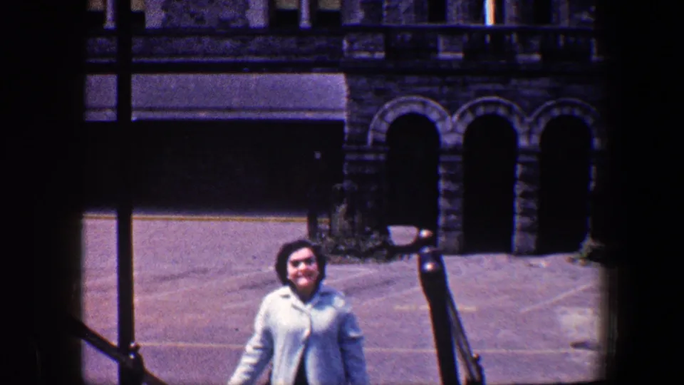 Late moment of the 1960 Baltimore shot, with the woman in a light blue coat on steps before arched stone doorways and a paved plaza.