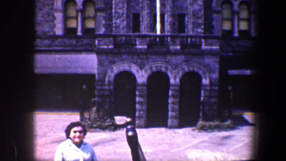 The woman remains posed outside the Baltimore stone building, with dark arches and a pole partly crossing the 8mm frame.