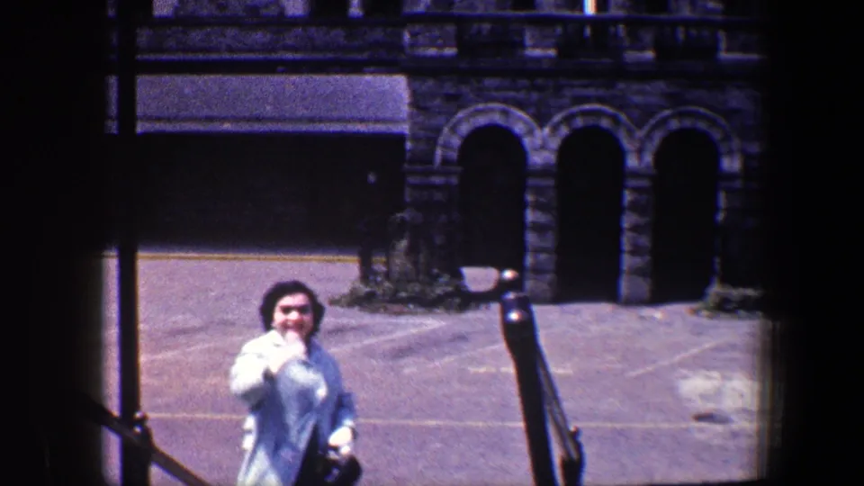In this 1960 Baltimore frame, the woman raises a hand toward the camera while standing before stone arches and a paved forecourt.