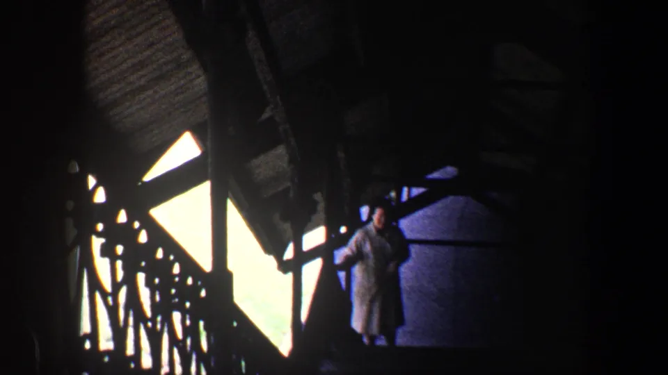 1960 Baltimore home-movie frame of a woman in a light grey coat on a shadowed stairway with strong window backlight.
