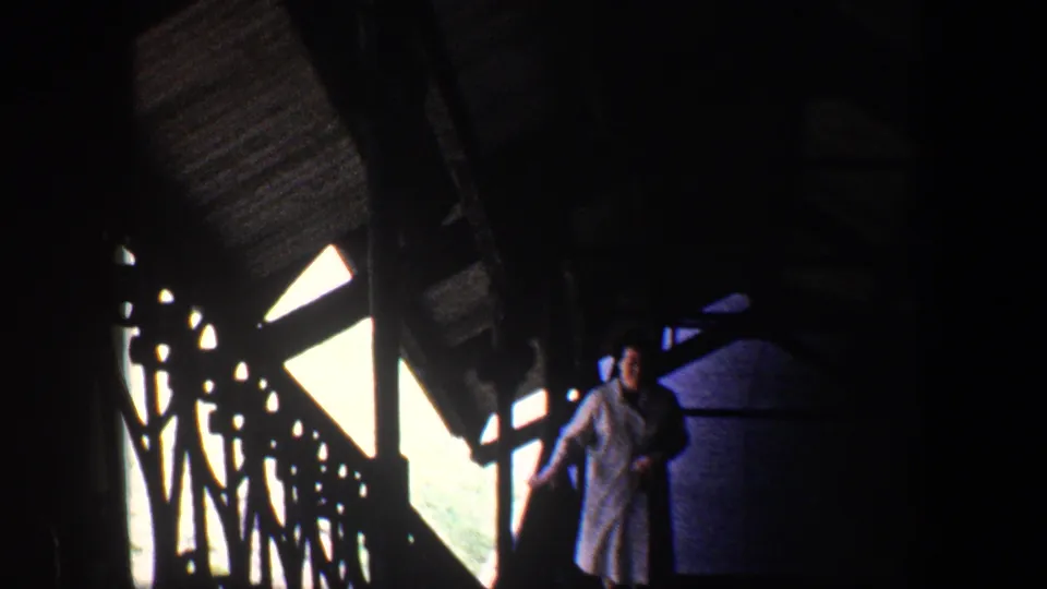 1960 Baltimore home-movie frame of a woman ascending a dim stairway, framed by railing silhouettes and bright window light.