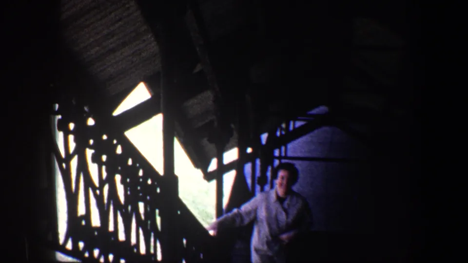 The woman smiles toward the camera while walking up a dim stairway, with strong window light and railing silhouettes visible around her.