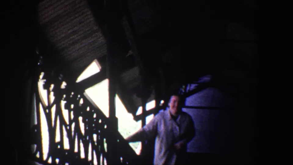 The woman comes closer on the staircase, smiling within a high-contrast 8mm interior scene of railings, shadows, and bright window openings.
