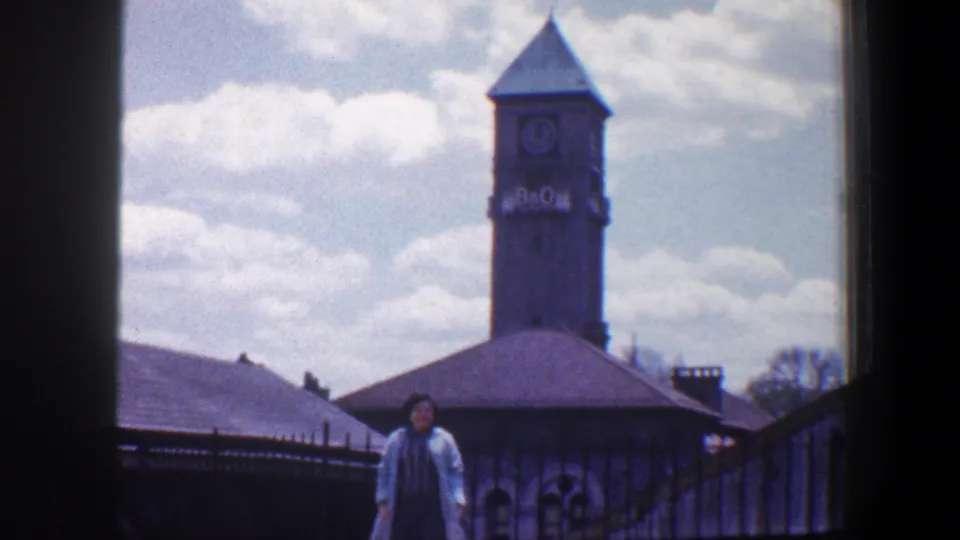 A woman poses outdoors near railings with the B&O clock tower and rooftops visible behind her in Baltimore, circa 1960.