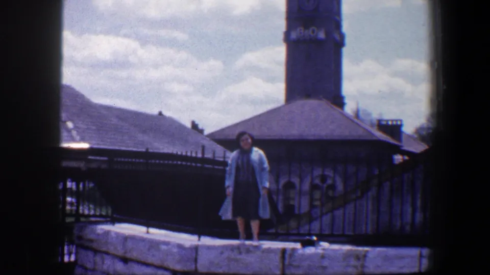 A woman in a light coat stands on a stone edge in front of railings, rooftops, and the B&O clock tower in Baltimore.