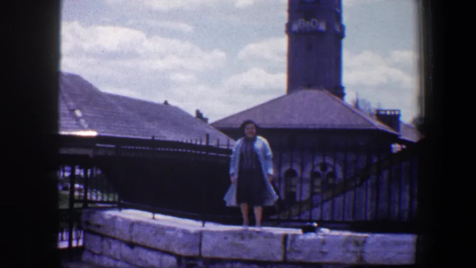 In 1960 Baltimore footage, a woman in a dress and light coat stands on a stone wall near iron fencing and a clock tower.