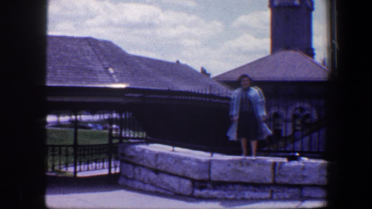The woman remains posed on the wall as the frame includes iron railings, a low roof, and the tower in the Baltimore setting.