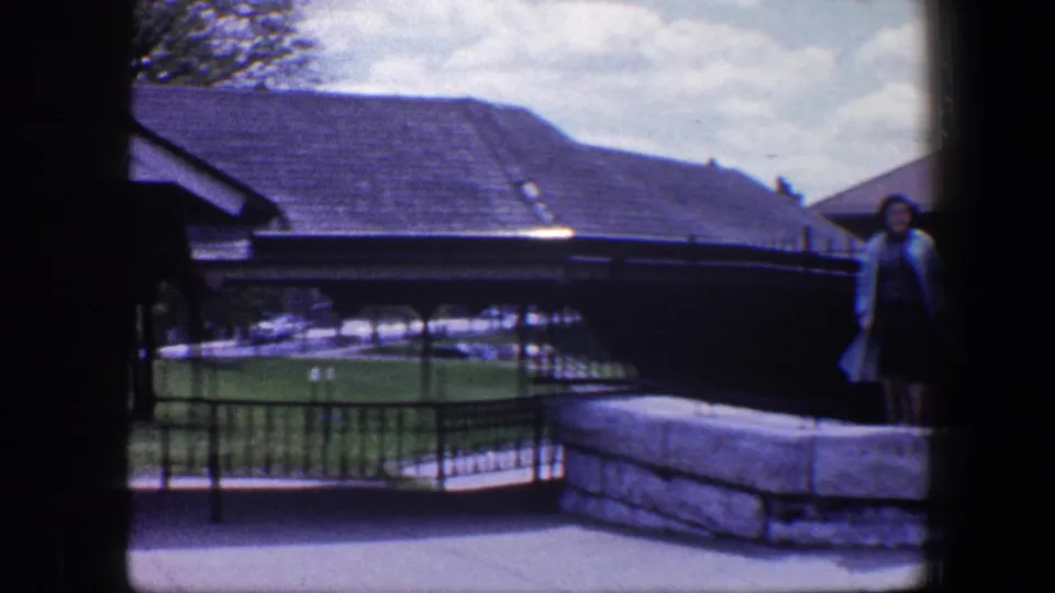The handheld pan shifts left, placing the woman at the edge while grass, iron fencing, rooflines, and stonework fill the frame.