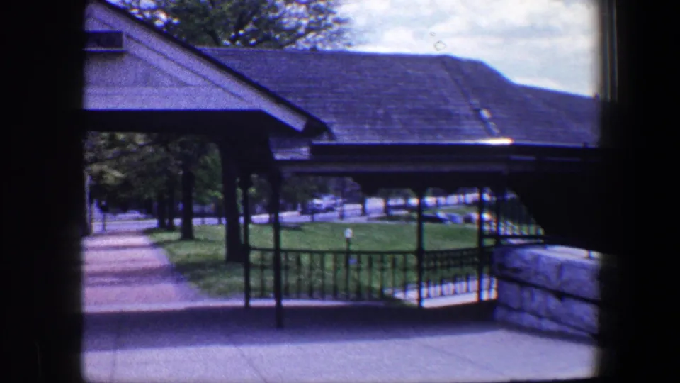 The final frame looks across the iron fence toward grass, trees, a walkway, and a roofed structure in the Baltimore exterior.
