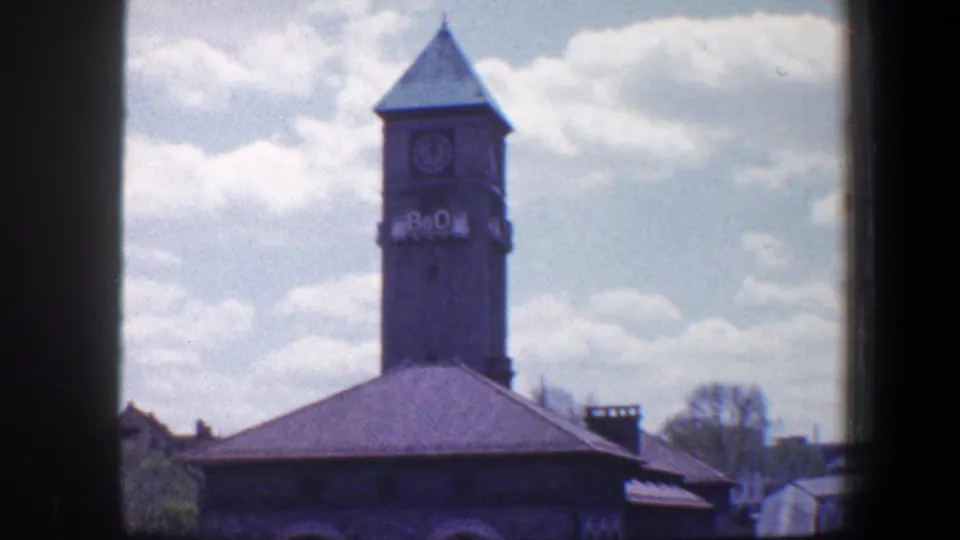 1960 Baltimore 8mm frame showing a grey stone clocktower with B&O lettering above a low roofline and cloudy sky.