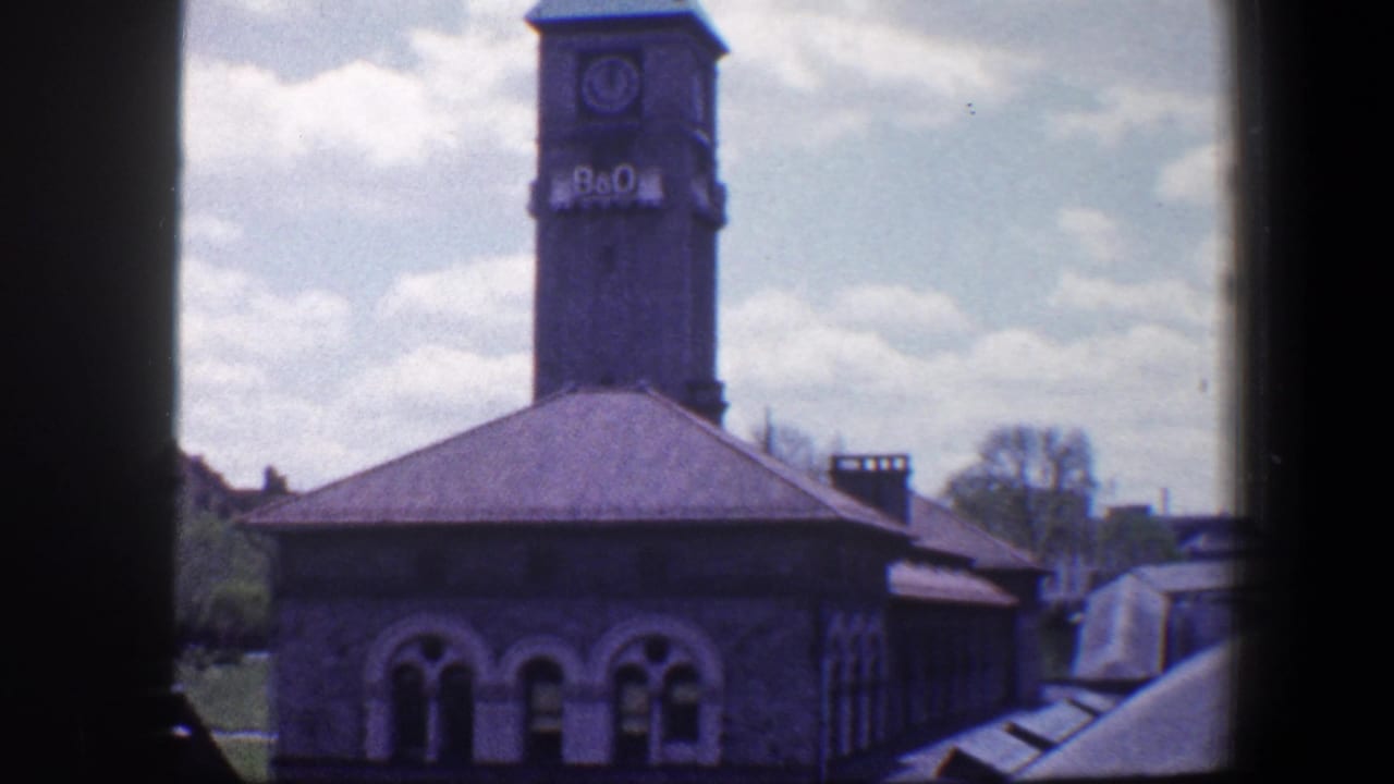 Baltimore 1960 home-movie view of the B&O clocktower above a stone building with arched windows and bright clouds.