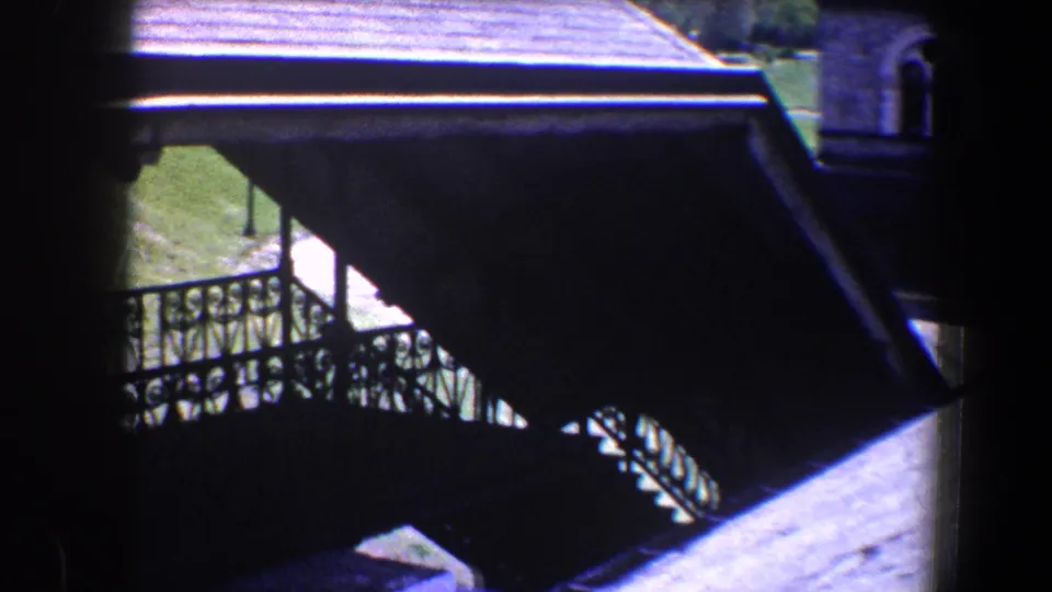 1960 Baltimore home-movie frame showing a covered exterior stair with iron railing between roofs and stone architecture.