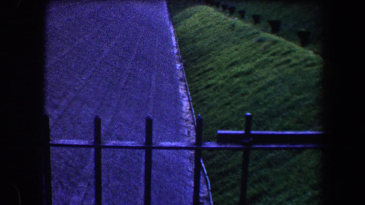 Balcony railing, paved path, stone wall, and grassy hillside seen in soft 8mm color.