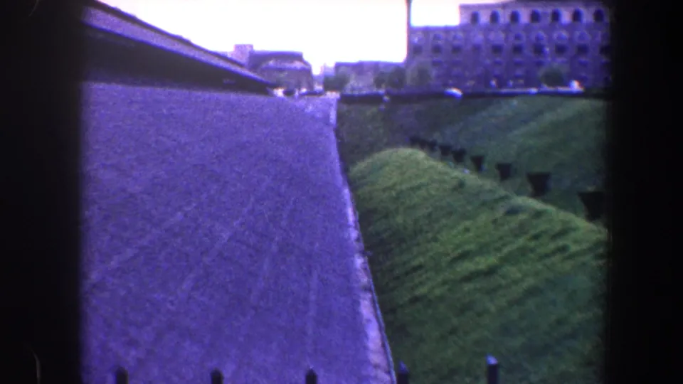 1960 Baltimore frame from a balcony showing a path, green hillside, railing posts, and a distant masonry building.