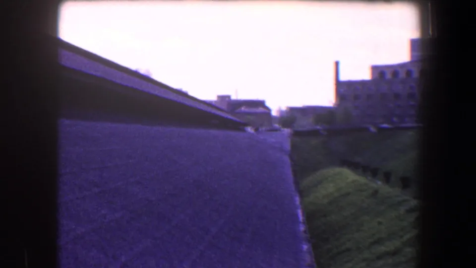 1960 Baltimore home-movie frame with a balcony wall, sloped grass, distant buildings, and bright overexposed sky.