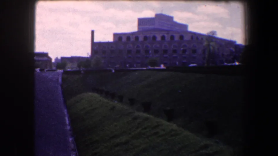 The 1960 home-movie view centers on a large arched industrial-looking building beside a road and grassy embankment in Baltimore.