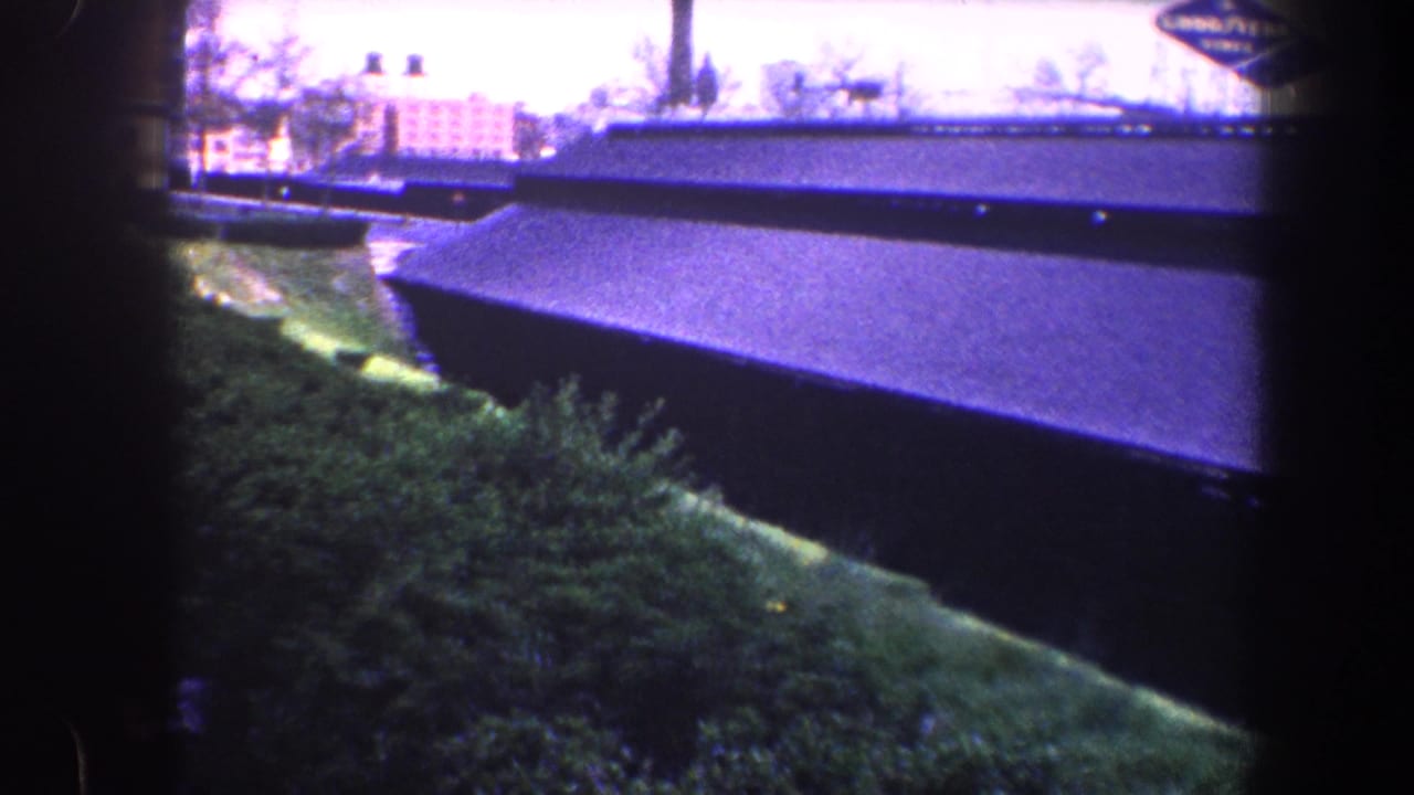 1960 Baltimore 8mm frame showing a long dark shed roof, green embankment, shrubs, and distant low buildings.