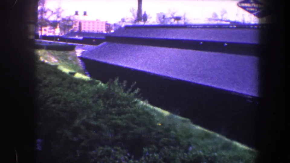 1960 Baltimore 8mm frame showing a long dark shed roof, green embankment, shrubs, and distant low buildings.