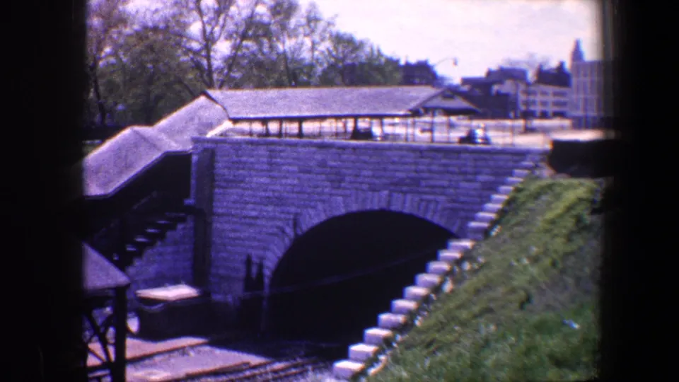 1960 Baltimore model railway layout with a stone tunnel, grassy slope, stairway, roofed platform, tracks, and trees behind it.