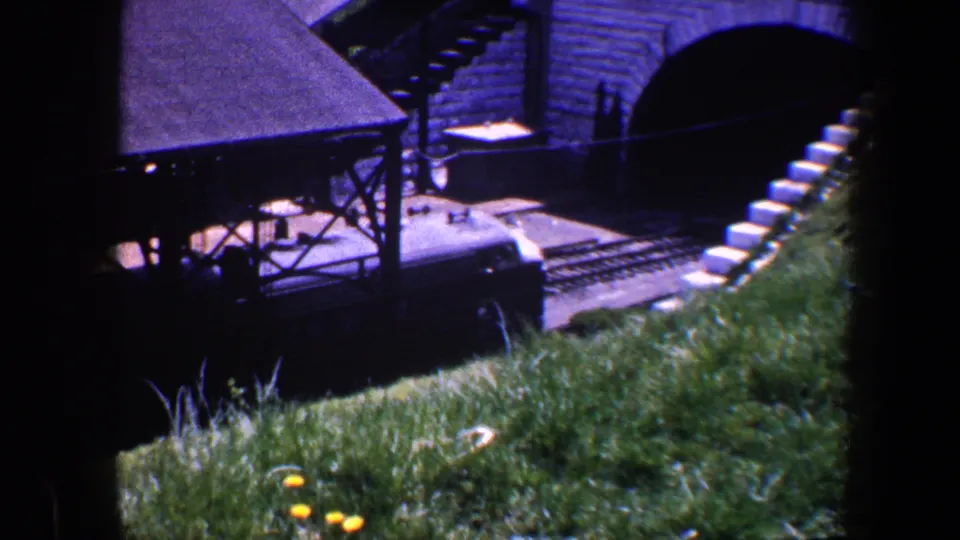 1960 Baltimore 8mm frame showing a dark train passing beneath a brick arched bridge, viewed from a grassy embankment.