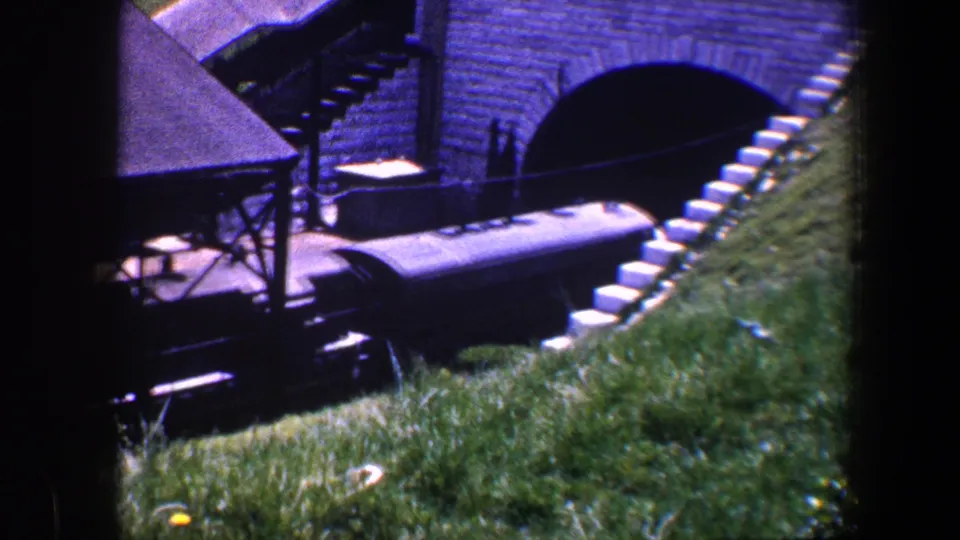 The train continues beneath the arched brick structure, with the sloped grass and trackside platform still visible.