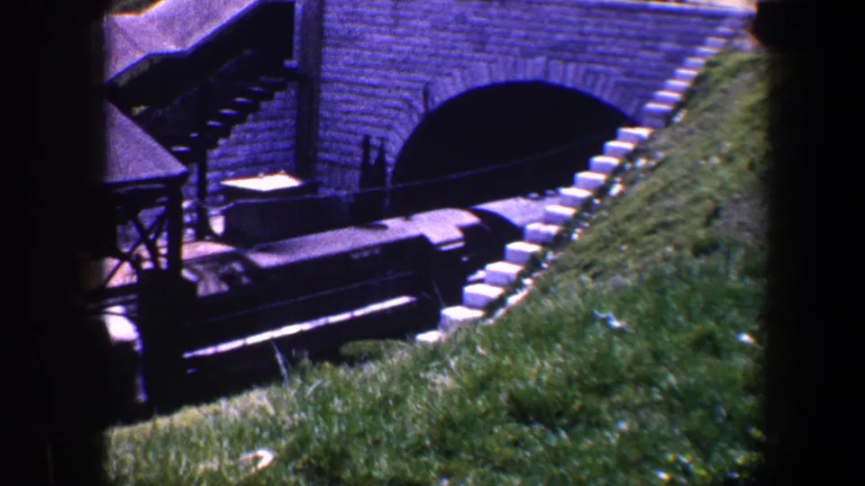 A longer section of the dark train is visible below the brick arch, framed by grass, rails, and the bridge structure.