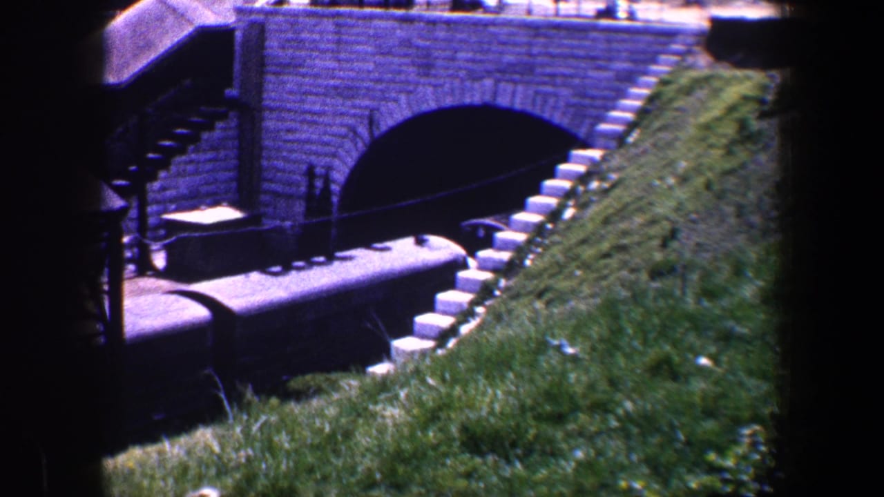 1960 Baltimore 8mm frame showing freight cars near a stone arch tunnel under brickwork, with grass and trackside structures visible.
