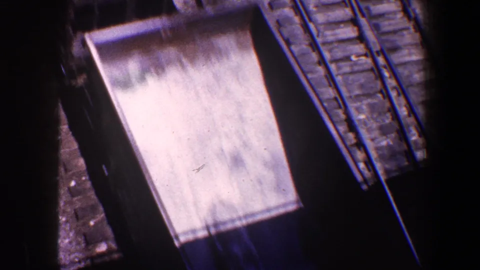 Close elevated 8mm view of rail tracks and a freight car roof near masonry work, with strong shadow and film gate edges.