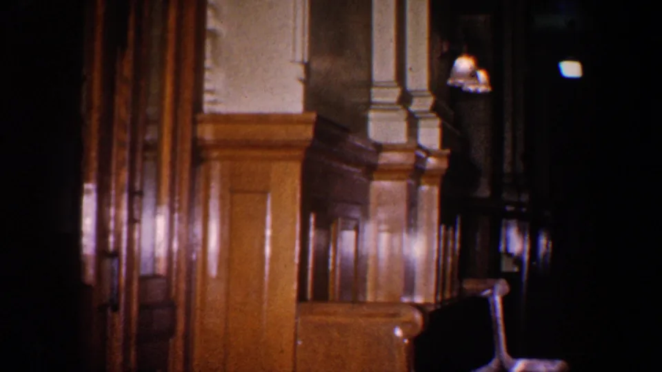 Brief 1960 8mm frame of a wood-paneled interior or corridor-like space with columns, a dark doorway, and warm reflected light.