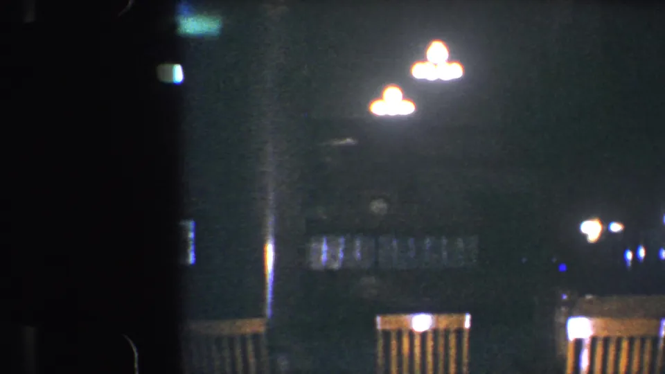 Baltimore 1960 8mm frame showing a dim empty dining space with warm lights, wooden chairs, and dark reflective surfaces.