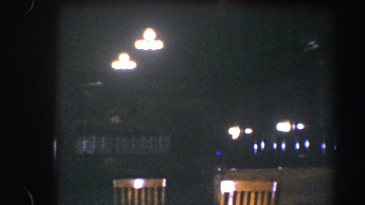 1960 Baltimore 8mm frame showing dim overhead lights, wooden chairs, and blurred background lamps in a dark setting.