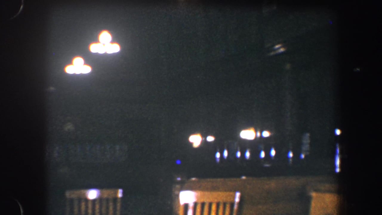 1960 Baltimore home-movie frame with bright overhead fixtures, empty chair backs, and soft points of light beyond them.