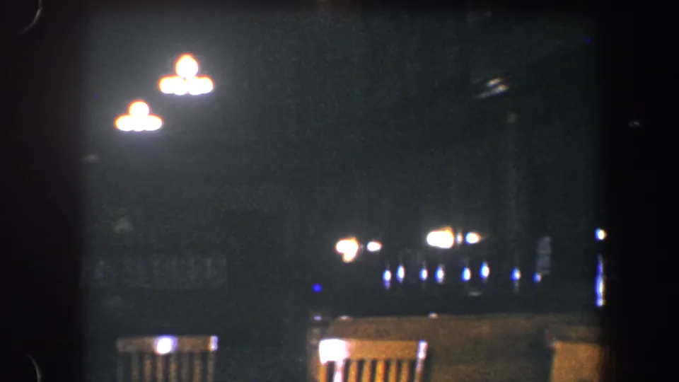 1960 Baltimore home-movie frame with bright overhead fixtures, empty chair backs, and soft points of light beyond them.