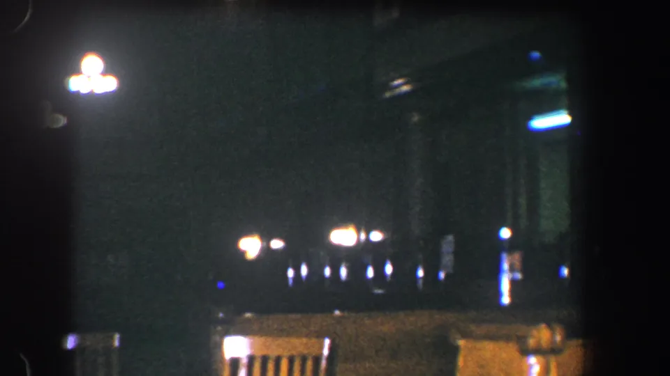 1960 Baltimore 8mm frame showing empty wooden chairs, scattered lamps, and a bluish light in a dim night environment.